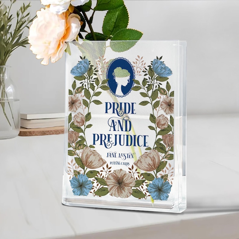 K&ouml;nnte beinhalten: Ein klarer Acryl-Spielkartenhalter mit dem Titel "Pride and Prejudice" von Jane Austen. Das Design umfasst einen Blumenkranz mit blauen und braunen Blumen und eine Silhouette einer Frau.