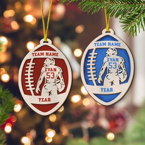 May include: Two wooden football ornaments with a player in the middle. The ornament on the left is red and says "Team Name", "Evan 53", and "Year". The ornament on the right is blue and says "Team Name", "Evan 53", and "Year".