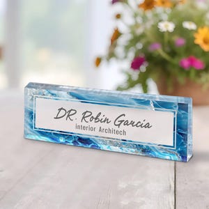 Può includere: Un portapiastrello da scrivania in vetro trasparente con un design astratto blu e bianco. Il portapiastrello riporta la scritta "DR. Robin Garcia, architetto d'interni"