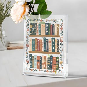 May include: Clear acrylic vase with a bookshelf design. The vase features a colorful illustration of books on shelves, surrounded by a floral border. The vase is rectangular and sits on a white surface.