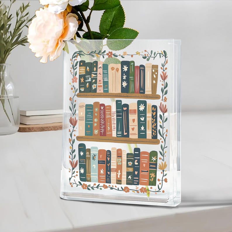 May include: Clear acrylic vase with a bookshelf design. The vase features a colorful illustration of books on shelves, surrounded by a floral border. The vase is rectangular and sits on a white surface.