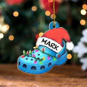 Puede incluir: Adorno navideño azul en forma de zueco con un gorro de Papá Noel rojo y el nombre "MARK" en blanco. El zueco está decorado con una guirnalda de luces navideñas de colores. Una decoración festiva.