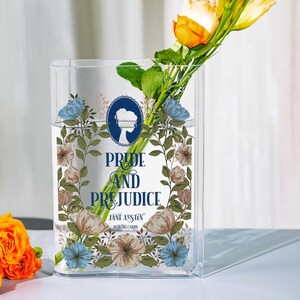 K&ouml;nnte beinhalten: Eine klare, rechteckige Glasvase mit dem Text "Pride and Prejudice" und "Jane Austen Playing Cards", umgeben von floralen Illustrationen. Die Vase enth&auml;lt Wasser und einen Strau&szlig; gelber und orangefarbener Blumen.