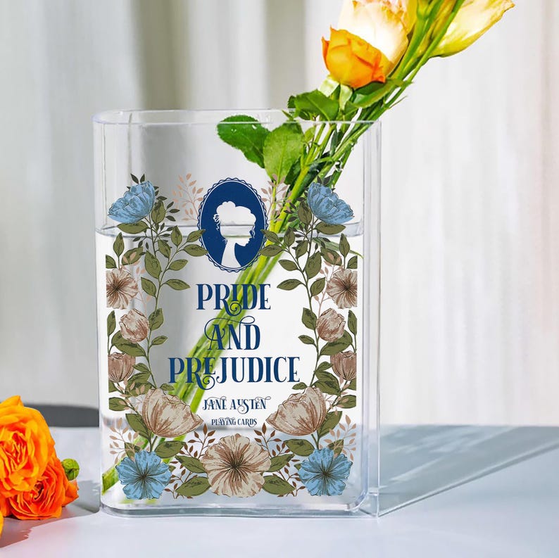 K&ouml;nnte beinhalten: Eine klare, rechteckige Glasvase mit dem Text "Pride and Prejudice" und "Jane Austen Playing Cards", umgeben von floralen Illustrationen. Die Vase enth&auml;lt Wasser und einen Strau&szlig; gelber und orangefarbener Blumen.