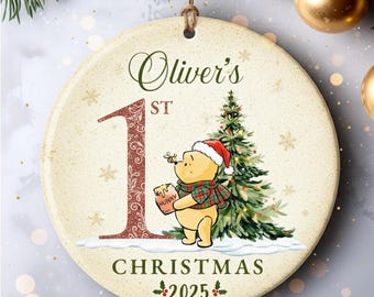 Op de afbeelding: Ronde kerstversiering met de tekst "Oliver's 1st Christmas 2025". Het heeft een cartoon illustratie van Winnie de Poeh met een kerstmuts en een honingpot. Het cijfer 1 is in een decoratief lettertype.