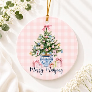 Può includere: Un ornamento rotondo in ceramica con sfondo a quadretti rosa chiaro che raffigura un albero di Natale verde in un vaso blu e bianco, decorato con tessere del Mahjong e palline, sormontato da un fiocco rosa e la scritta Merry Mahjong.