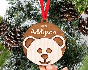May include: A round wooden Christmas ornament featuring a light-colored wood bear face cutout layered over a dark wood circular base. The ornament displays the name Addyson and the year 2024 in white text, suspended by a bright red ribbon.