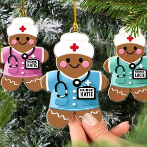 Könnte beinhalten: Drei Lebkuchen-Krankenschwester-Ornamente mit goldenen Aufhängeschnüren. Jedes Ornament zeigt einen lächelnden Lebkuchenmann in einer Krankenschwesteruniform, komplett mit Stethoskop und Namensschild mit der Aufschrift "Nurse Katie" oder "Nurse Lucie". Die Uniformen sind rosa, blau und türkis.