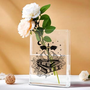 Pode incluir: Um vaso de vidro transparente e retangular com uma ilustração preta de livros empilhados, um dragão e estrelas. O vaso contém água e um caule de rosas brancas e pêssego com folhas verdes. Uma bola decorativa e uma rosa estão sobre a mesa.