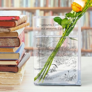 Puede incluir: Un jarr&oacute;n de vidrio transparente y rectangular con una ilustraci&oacute;n y cita de Winnie the Pooh, que contiene rosas amarillas y naranjas. Una pila de libros antiguos est&aacute; a la izquierda. El jarr&oacute;n est&aacute; lleno de agua.