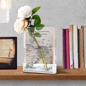 Puede incluir: Un jarr&oacute;n rectangular de vidrio transparente con una ilustraci&oacute;n de Winnie the Pooh y la cita: "Sometimes, I sits and thinks and sometimes, I just sits." El jarr&oacute;n contiene un tallo de rosas con flores blancas y color melocot&oacute;n. Una imagen enmarcada y libros est&aacute;n en el fondo.