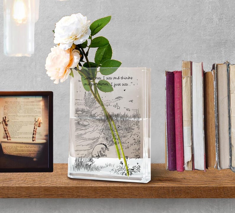 Puede incluir: Un jarr&oacute;n rectangular de vidrio transparente con una ilustraci&oacute;n de Winnie the Pooh y la cita: "Sometimes, I sits and thinks and sometimes, I just sits." El jarr&oacute;n contiene un tallo de rosas con flores blancas y color melocot&oacute;n. Una imagen enmarcada y libros est&aacute;n en el fondo.