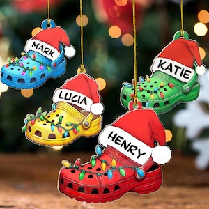 Puede incluir: Cuatro adornos navideños con forma de zapatos Crocs, cada uno de un color diferente: azul, verde, amarillo y rojo. Cada zapato tiene un gorro de Papá Noel y una guirnalda de luces de colores. Los nombres están impresos en los gorros: Mark, Katie, Lucia y Henry.