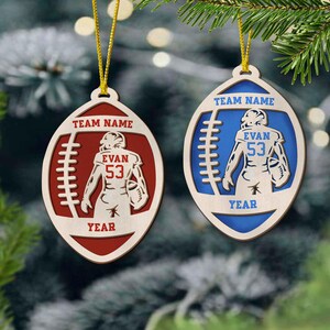 May include: Two wooden football ornaments with a player in the middle. The ornament on the left is red and the ornament on the right is blue. Both ornaments have the text "TEAM NAME", "EVAN 53", and "YEAR".