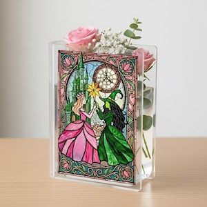 Puede incluir: Jarrón de acrílico transparente con un panel de vidriera que representa a una princesa y una bruja sosteniendo una estrella. El panel tiene un castillo, un reloj y un borde floral. Rosas rosas y vegetación están dentro del jarrón.