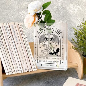 May include: A clear acrylic bookend with the text "A Book A Day keeps the mental breakdown away" and a fantasy illustration. A row of books and a vase with flowers are on a wooden shelf.