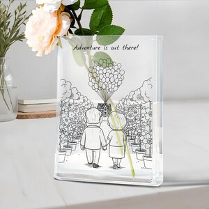 Può includere: Vaso rettangolare trasparente con un'illustrazione in bianco e nero di una coppia che cammina verso una casa sollevata da palloncini. Il testo "Adventure is out there!" è sopra l'illustrazione. Un singolo fiore e del verde sono all'interno del vaso.