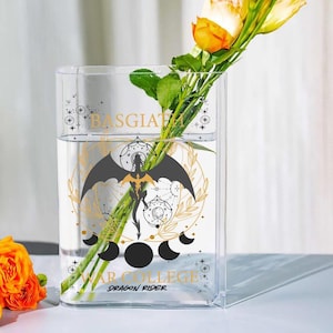 May include: A clear rectangular glass vase with water and yellow roses. The vase features a black and gold dragon design with the text "BASGIATH" and "WAR COLLEGE DRAGON RIDER". The vase is on a white surface.