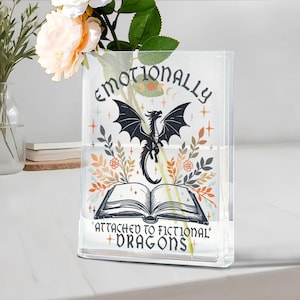 May include: Clear glass vase with the text "Emotionally Attached to Fictional Dragons" and a black dragon illustration. The vase has a rectangular shape and is filled with water. Floral accents surround the dragon and open book design.