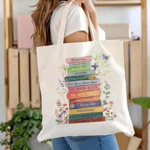 Może przedstawiać: Kremowa torba typu tote z nadrukiem stosu kolorowych grzbietów książek, w tym "Harry Potter" i "The Cruel Prince." Kwiatowe akcenty otaczają stos książek. Torba jest trzymana przez osobę.