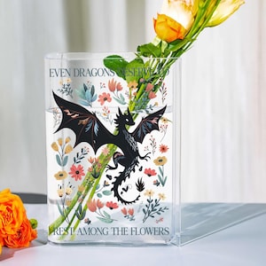 May include: A clear rectangular vase with a black dragon illustration and floral accents. The vase contains water and yellow roses. Text on the vase reads "EVEN DRAGONS DESERVE TO REST AMONG THE FLOWERS."