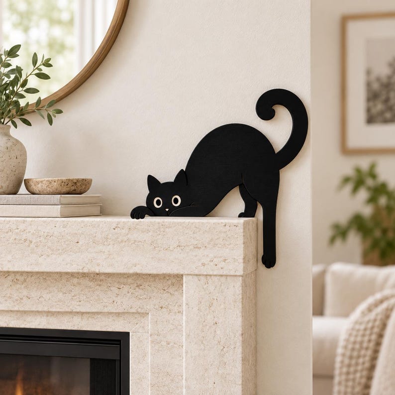 May include: A flat, black silhouette of a cat with large white eyes and a curled tail, designed to be positioned on the edge of a surface. The cat is depicted in a crouching posture with its front paws extended over the edge of a mantelpiece.