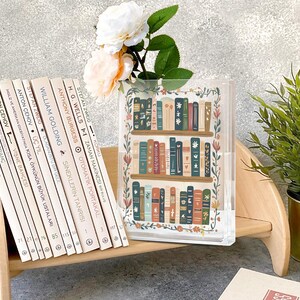 May include: A clear acrylic book vase with a colorful illustration of a bookshelf filled with books. The vase is decorated with floral accents and holds white and peach flowers. A row of books in Turkish is on a wooden shelf.