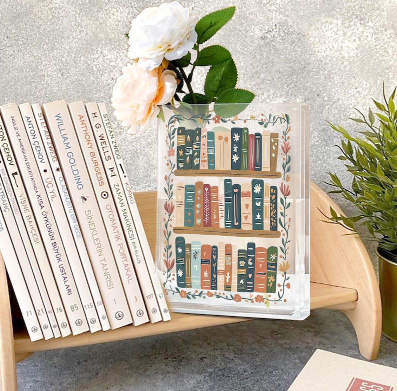 May include: A clear acrylic book vase with a colorful illustration of a bookshelf filled with books. The vase is decorated with floral accents and holds white and peach flowers. A row of books in Turkish is on a wooden shelf.