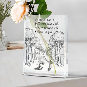 May include: Rectangular clear glass vase with a black and white illustration of Winnie the Pooh and Piglet in a forest scene. The vase displays the quote, "It makes such a Difference said Pooh to have someone who Believes in you."