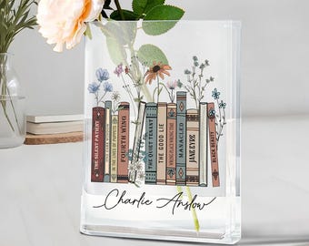 Puede incluir: Un jarrón de cristal transparente con una fila de lomos de libros ilustrados, incluyendo títulos como "The Silent Patient" y "Jane Eyre". El jarrón está personalizado con el nombre "Charlie Anslow" y contiene flores. Un artículo decorativo para el hogar.