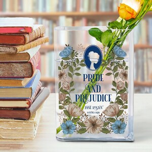 K&ouml;nnte beinhalten: Eine transparente, rechteckige Glasvase mit dem Text "Pride and Prejudice" und floralen Illustrationen. Die Vase enth&auml;lt Wasser und einen Strau&szlig; gelber und orangefarbener Rosen. Links ist ein Stapel B&uuml;cher zu sehen.