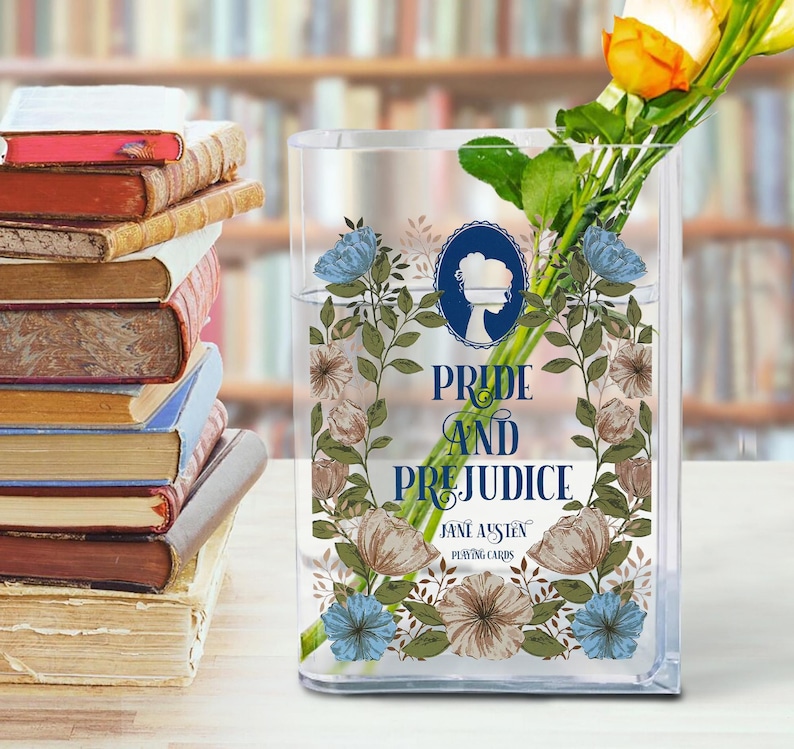 K&ouml;nnte beinhalten: Eine transparente, rechteckige Glasvase mit dem Text "Pride and Prejudice" und floralen Illustrationen. Die Vase enth&auml;lt Wasser und einen Strau&szlig; gelber und orangefarbener Rosen. Links ist ein Stapel B&uuml;cher zu sehen.