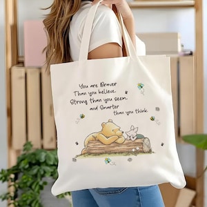 May include: Cream-colored tote bag featuring a cartoon illustration of Winnie the Pooh and Piglet on a log, with bees and the text "You are Braver Than you believe. Strong than you seem. and Smarter than you think."