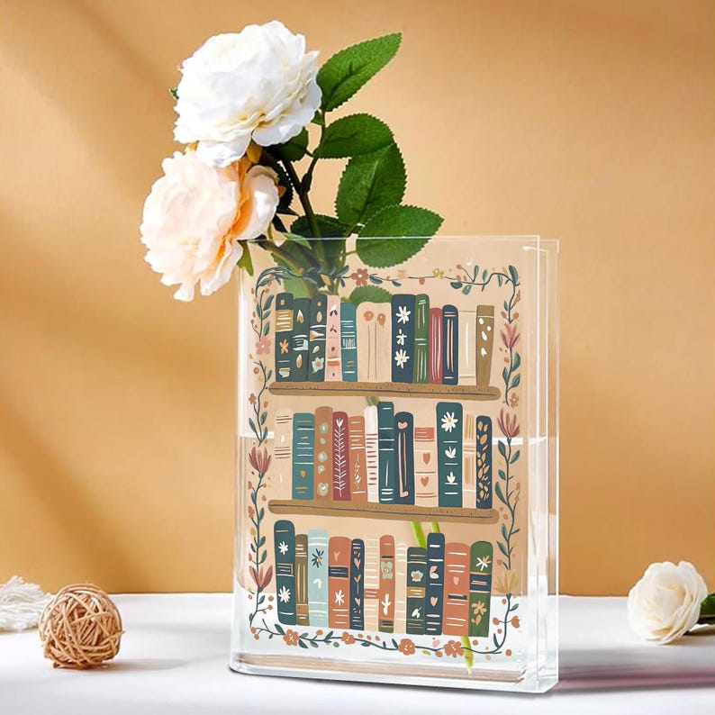 May include: Clear rectangular vase with a bookshelf design, filled with water and white and peach-colored flowers. The vase has a floral border and a decorative bookshelf illustration with colorful books.