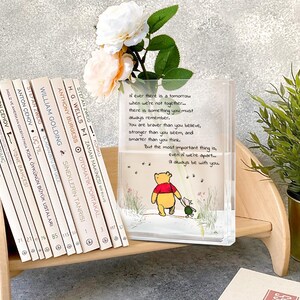 May include: A clear, rectangular vase with a Winnie the Pooh illustration and a quote. The vase holds white and peach flowers. Books with white spines are on a wooden shelf next to the vase.