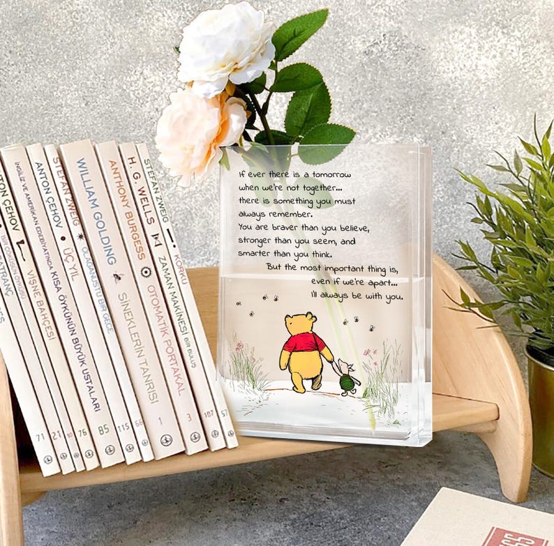 May include: A clear, rectangular vase with a Winnie the Pooh illustration and a quote. The vase holds white and peach flowers. Books with white spines are on a wooden shelf next to the vase.