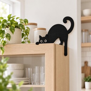 May include: A black cat-shaped decorative figure with a curved tail and white eyes, designed to perch on the edge of a shelf or cabinet. The flat, silhouette-style ornament features a matte finish and is positioned with its front paws resting over the top corner of a light-colored wooden furniture piece.