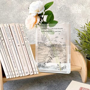 Puede incluir: Un jarr&oacute;n rectangular transparente con una ilustraci&oacute;n de Winnie the Pooh y la cita, "Sometimes, I sits and thinks and sometimes, I just sits..." est&aacute; lleno de agua y flores. Una pila de libros est&aacute; a la izquierda.