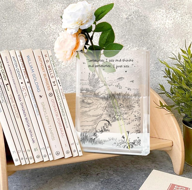 Puede incluir: Un jarr&oacute;n rectangular transparente con una ilustraci&oacute;n de Winnie the Pooh y la cita, "Sometimes, I sits and thinks and sometimes, I just sits..." est&aacute; lleno de agua y flores. Una pila de libros est&aacute; a la izquierda.