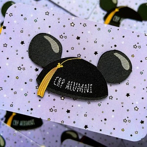 May include: A black and yellow embroidered patch featuring a Mickey Mouse ear hat design. The hat has a graduation cap on top with a tassel and the text "Cap Alumni". The patch is on a purple background with white stars.
