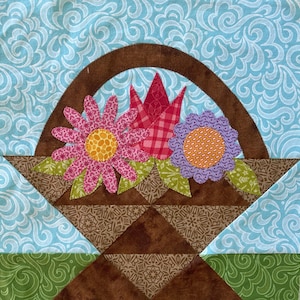 Könnte beinhalten: Ein brauner Stoffkorb mit einem gebogenen Griff, gefüllt mit rosa, roten und violetten Stoffblumen. Der Korb befindet sich auf einem blauen und grünen Stoffhintergrund.