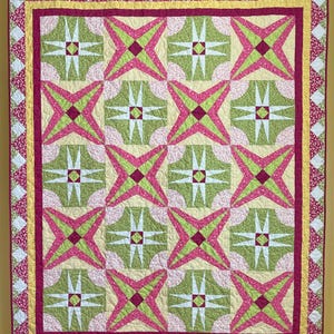 May include: A pink, green and white quilt with a geometric pattern. The quilt is hanging on a wall with a yellow background.