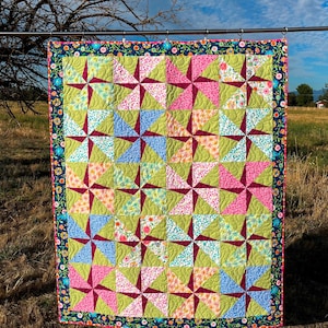 May include: A handmade quilt with a pinwheel design. The quilt features squares of green, pink, blue, and floral patterned fabric, with a dark red center. The border is a dark blue with a floral pattern. The quilt is hanging outside.