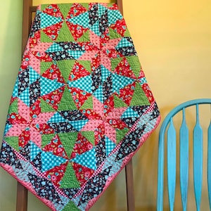 May include: A patchwork quilt with a geometric pattern in shades of pink, red, green, blue, and white. The quilt is hanging on a wooden ladder against a yellow wall.