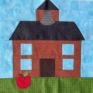 Może przedstawiać: Blok materiału przedstawiający budynek szkoły z czerwono-brązowym korpusem, czarnym dachem i jasnoniebieskimi oknami. Czerwone jabłko z zielonym liściem znajduje się na zielonej podstawie z tkaniny, z jasnoniebieskim tłem nieba.