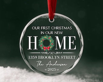 Pode incluir: Um enfeite de Natal redondo de vidro transparente pendurado por uma fita vermelha. O texto diz: OUR FIRST CHRISTMAS IN OUR NEW HOME, 1359 BROOKLYN STREET, The Anderson, 2025. Uma guirlanda verde com um laço vermelho substitui a letra O de HOME.