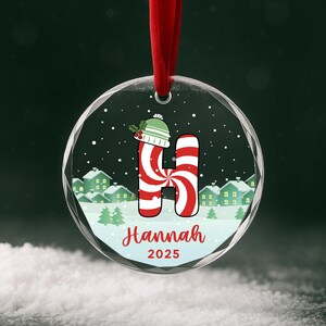 Pode incluir: Uma bola de Natal redonda com uma fita vermelha. Apresenta a letra "H" listrada de bengala doce, com um chapéu verde e branco. O nome "Hannah" e o ano "2025" estão escritos abaixo. Uma cena de inverno com neve e casas ao fundo.