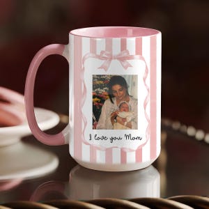 Puede incluir: Taza de cerámica blanca con rayas rosas y asa rosa. La taza presenta una foto enmarcada de una madre sosteniendo a un bebé, con un diseño de lazo rosa. El texto "I love you Mom" está escrito debajo.