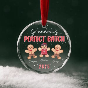 Puede incluir: Una bola de Navidad redonda y transparente con una cinta roja. El adorno presenta el texto "Grandma's PERFECT BATCH" en rojo y tres hombres de jengibre. También se incluyen nombres y el año 2025.