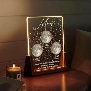 May include: A personalized night light with a dark background, featuring three moon images and the names Olivia, Alan, and Beth. The text reads "Mom" and "This is what the Moon looked like the day you became our Mom." The wooden base is illuminated.